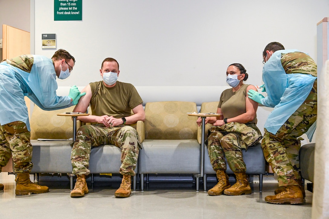 Two soldiers sit with their sleeves rolled up as two other service members administer shots to them.