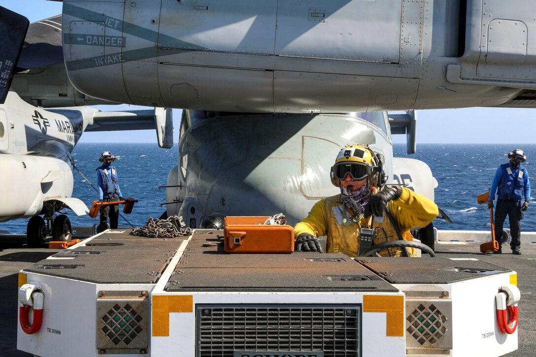 A sailor in yellow steers a vehicle moving an aircraft on a ship's deck.