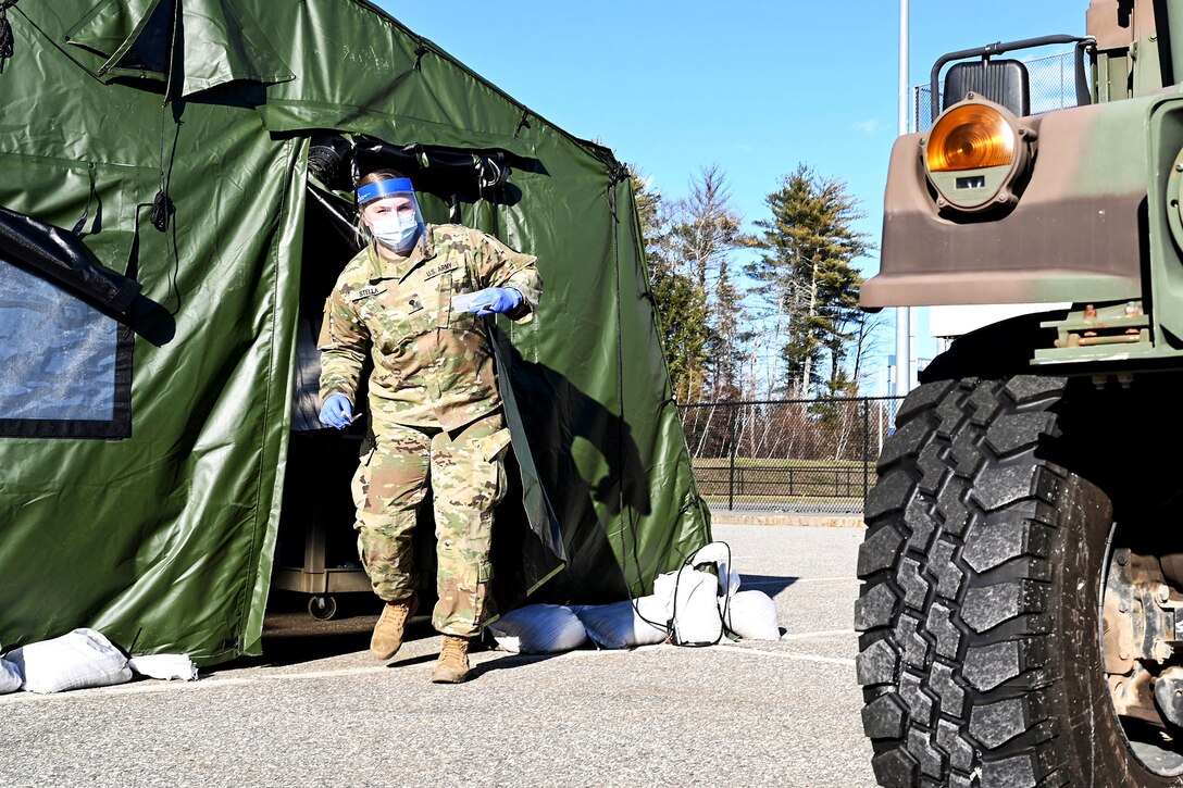 A soldier in personal protective equipment walks out of a green tent toward a vehicle in a paved lot.