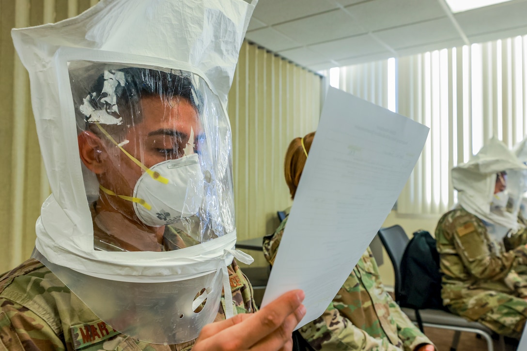 An airman sits with a group of other service members, all wearing face masks and hoods.