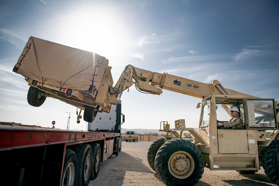 A soldier operates a forklift lifting a lare piece of equipment onto the back of a flatbed truck.