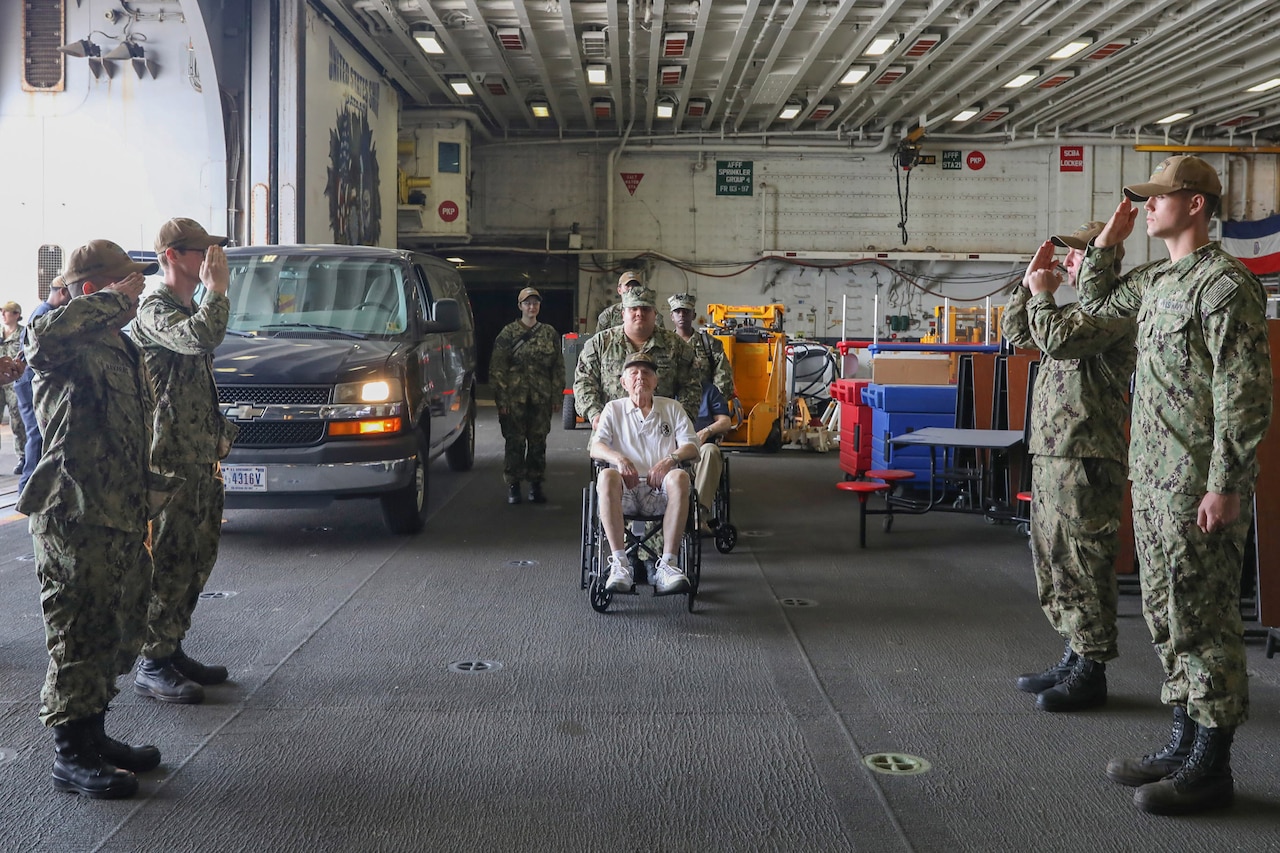 A man in a wheelchair is pushed forward. To his left and right, service members dressed in military uniforms salute.