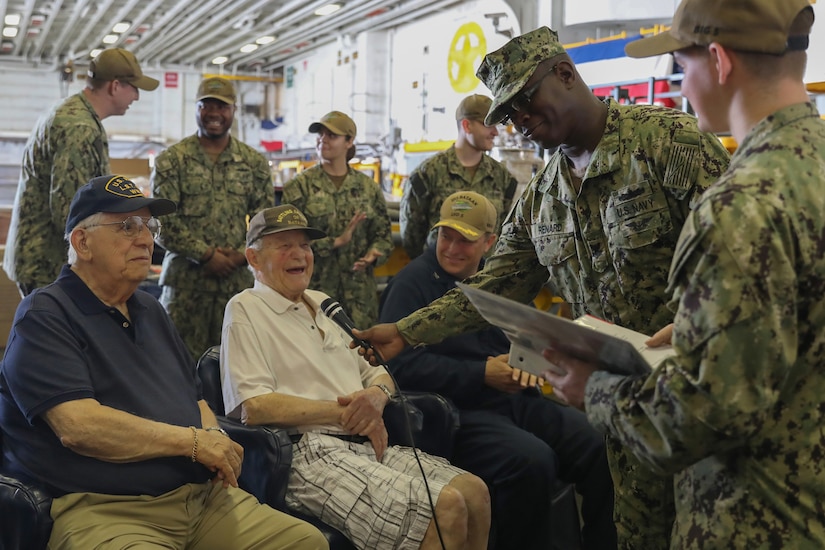 Several men are seated in chairs. They are surrounded by other individuals in military uniforms.