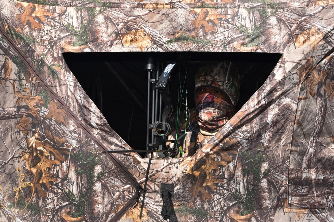 An airman wearing camouflage sits in a camouflage tent while holding a weapon.