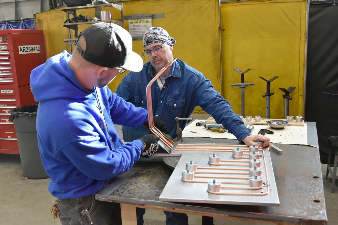 Blake Rich, left, and Darrell Hargrove, pipefitters in the Model and Machine Shop at Arnold Air Force Base, use a jig to bend copper tubing to fit within tape reel covers that will be installed in the 16-foot supersonic wind tunnel, known as 16S, at Arnold. This is among numerous efforts undertaken by craftsmen at Arnold which have contributed to the 16S return to service project. (U.S. Air Force photo by Bradley Hicks) (Image has been altered by obscuring badges for security purposes.)