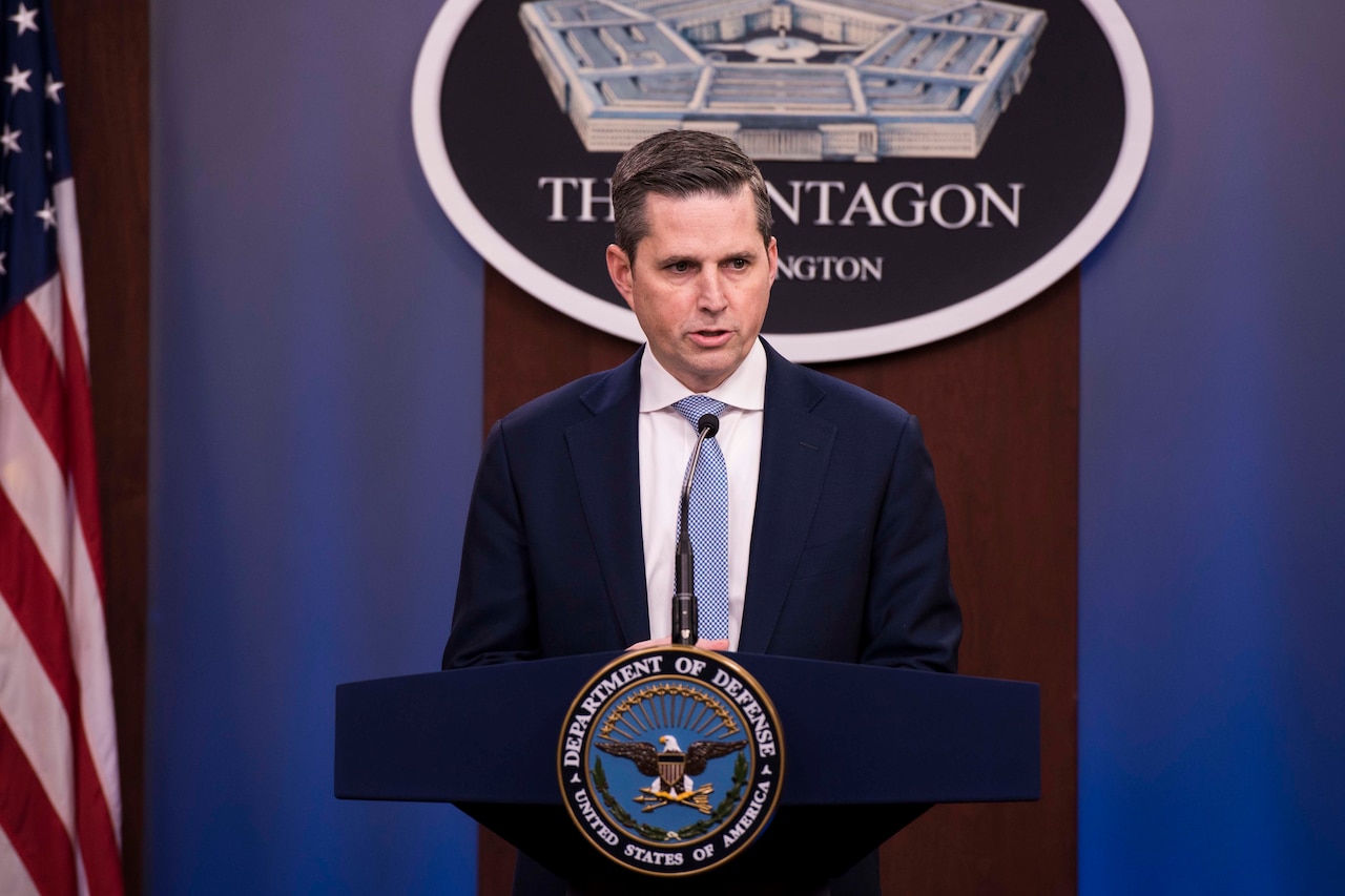 A man standing next to an American flag and in front of a Pentagon plaque speaks from a podium.