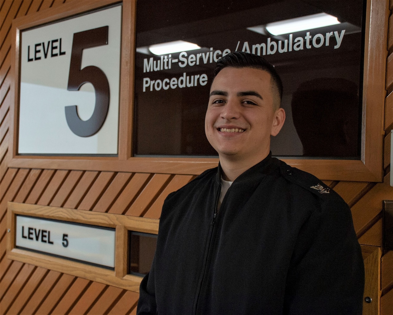 A sailor stands in front of a hospital unit sign in a hallway, smiling for the camera.