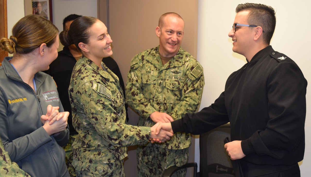 A sailor in a black jacket shakes the hand of another sailor in a Navy working uniform. Three other sailors stand around them smiling.
