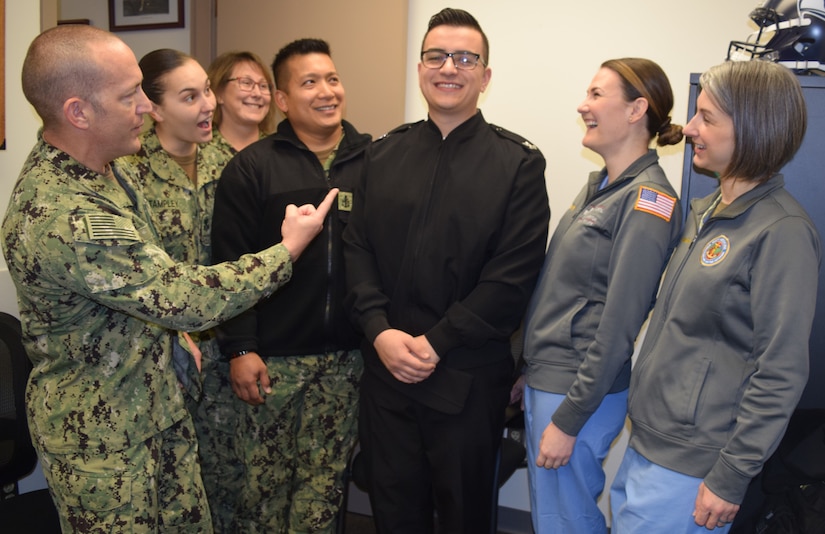 Seven Navy sailors laugh together in an office. One points toward the man in the middle.
