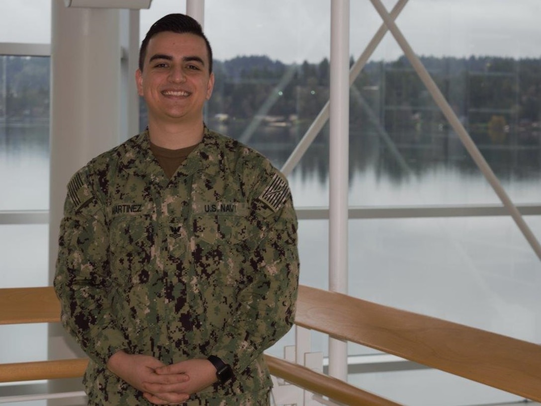 A sailor in his Navy working uniform poses for a photo by a window that overlooks a river.