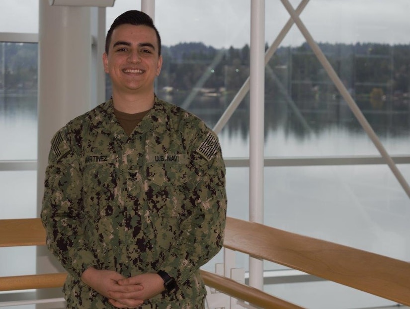 A sailor in his Navy working uniform poses for a photo by a window that overlooks a river.