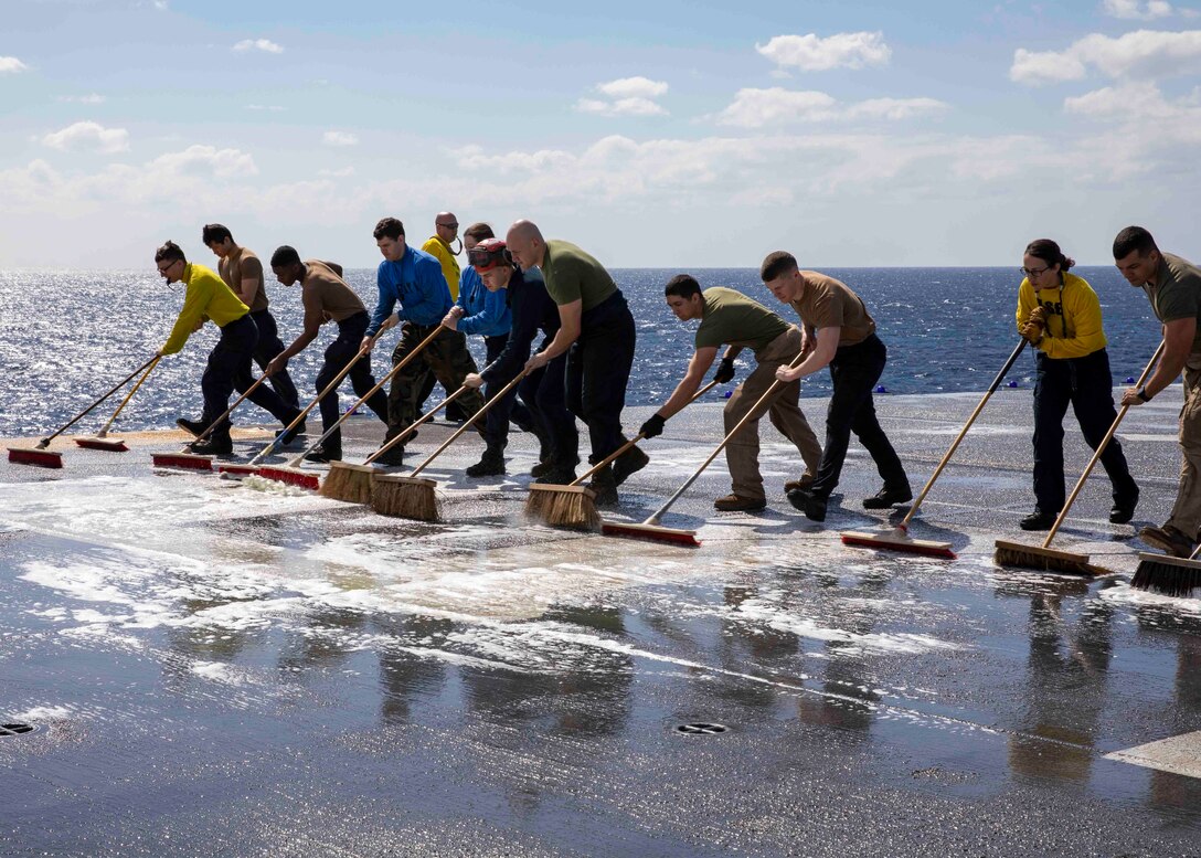 Sailors and Marines scrub a ship deck with brooms and water.