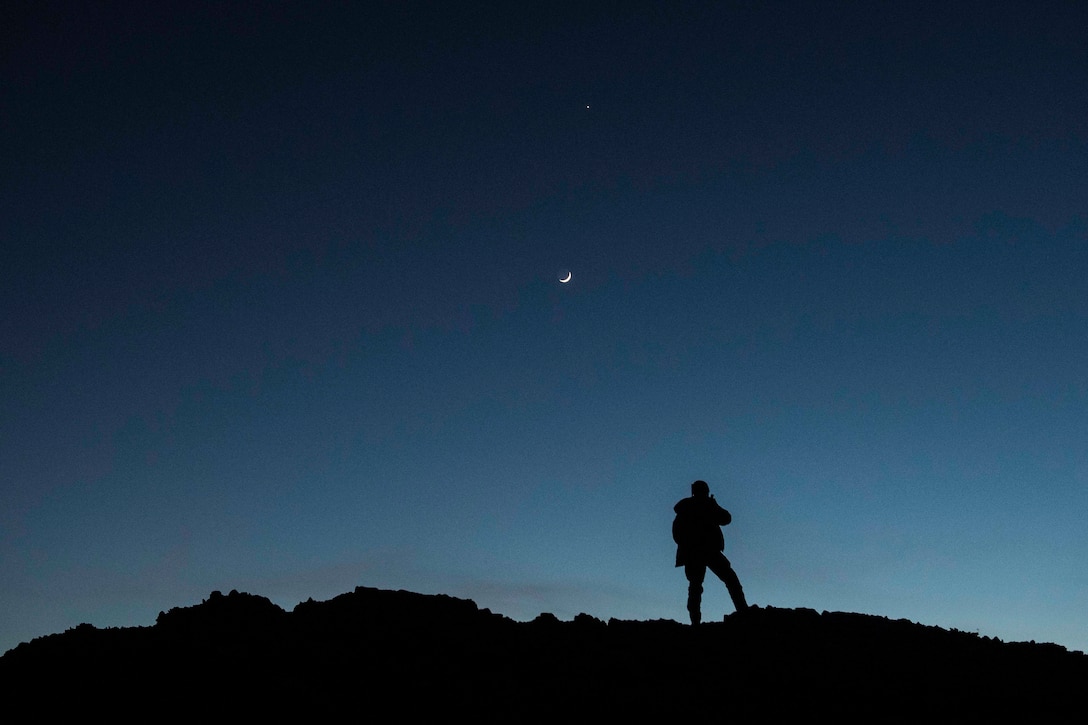 A silhouette of an airman standing at night; a crescent moon seen in the sky.