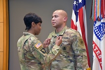 Female Soldier in green camouflage pins green and white ribbon on male Soldier's green camouflage uniform.