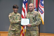 Female Soldier in green camouflage uniform holds green folder with white certificate with male Soldier in green camouflage uniform in front of the U.S. flag and the Army and Recruiting Command colors all of which are red, white and blue.