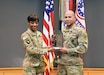 Female Soldier in green camouflage uniform holds bronze and black Soldier statue with male Soldier in green camouflage uniform in front of the U.S. flag and the Army and Recruiting Command colors all of which are red, white and blue.