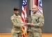 Female Soldier in green camouflage uniform holds bronze and black Soldier statue with male Soldier in green camouflage uniform in front of the U.S. flag and the Army and Recruiting Command colors all of which are red, white and blue.