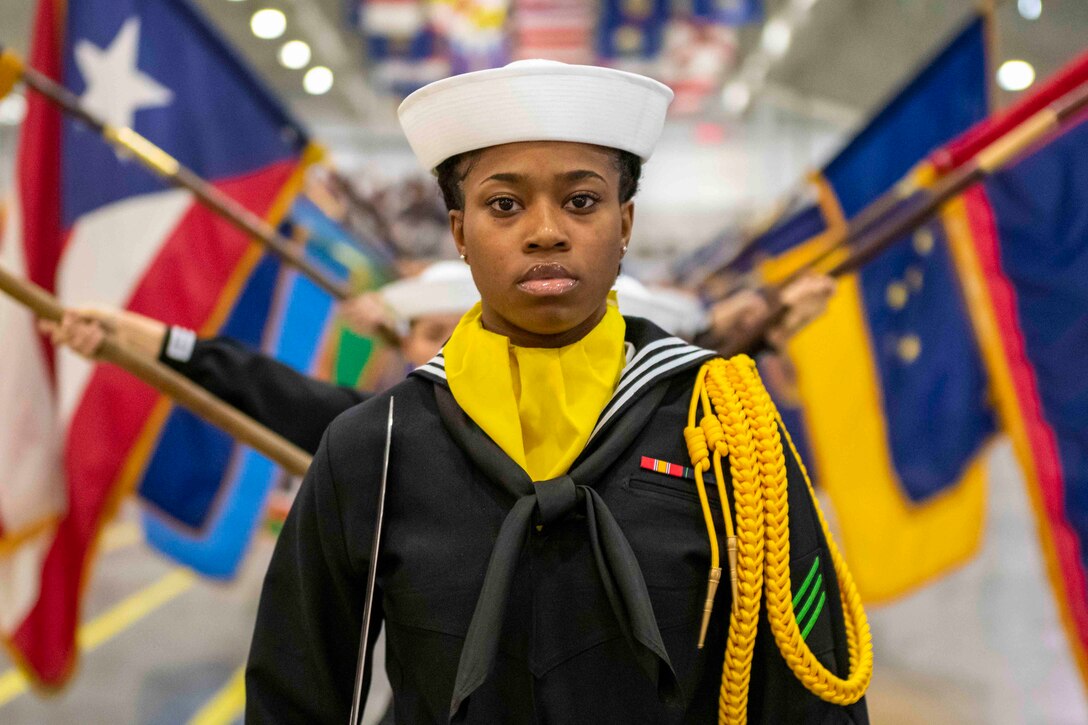 A Navy recruits walks in a line in front of people holding flags on either side.