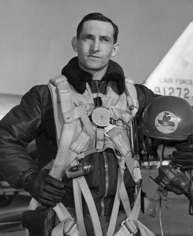 A pilot holds his parachute gear in one hand and his helmet in the other.