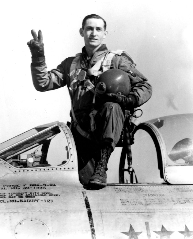A pilot stands in the cockpit of a plane with one leg propped on the side of it. He holds his helmet in one hand while forming a “V for Victory” sign with the fingers of his other hand.