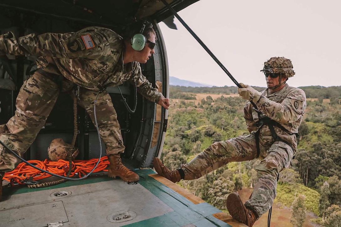 A soldier hangs off the side of a helicopter door while another soldier looks in his direction.