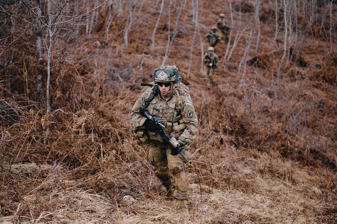 A soldier in uniform carries a weapon in the woods while other soldiers trail behind.