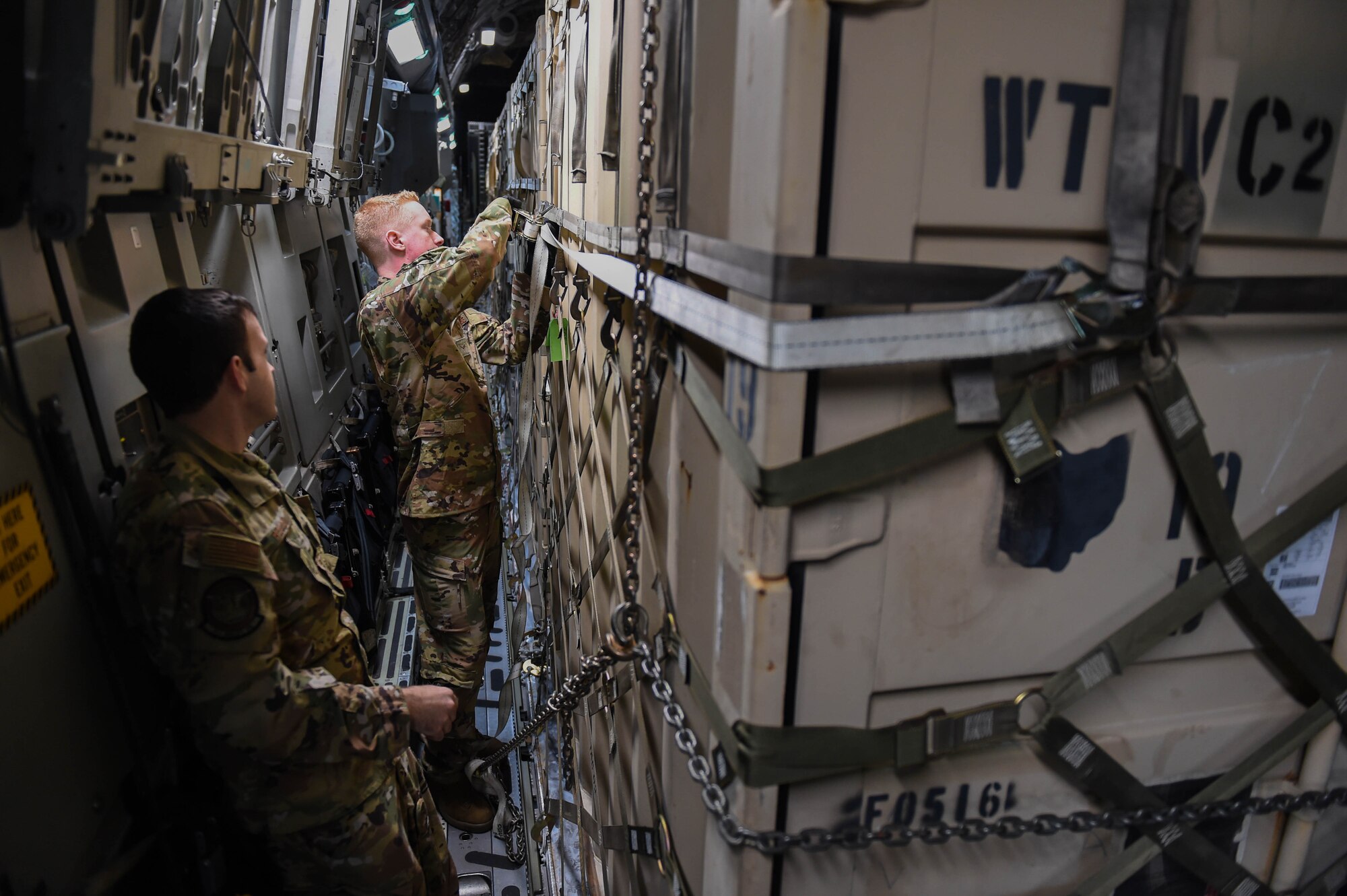 Master Sgt. Andrew Reilly, 4th Airlift Squadron loadmaster, observes Airman 1st Class Kam Watt, 4th Airlift Squadron loadmaster, attach and tighten an additional strap onto a piece of cargo aboard a C-17 Globemaster III before taking off from Spangdahlem Air Base, Germany, Dec. 18, 2019. The metal piece at the end of an existing strap had started to deteriorate, so as an added security measure the loadmasters attached one of their straps. (U.S. Air Force photo by Airman 1st Class Mikayla Heineck)