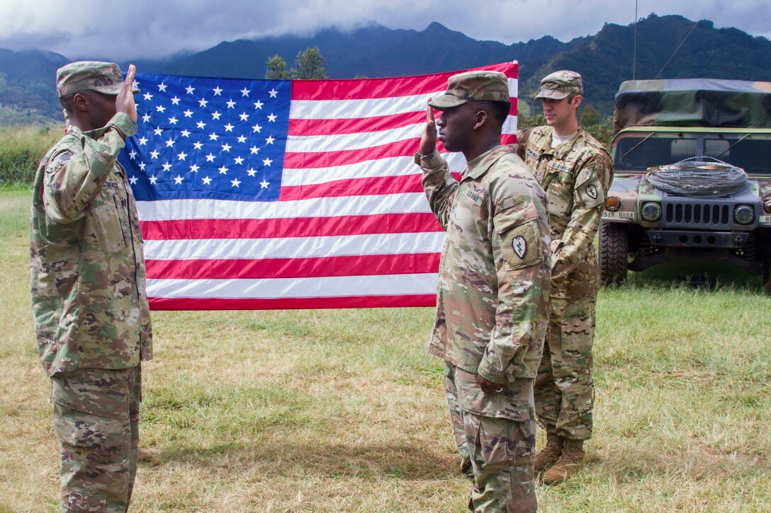 Two soldiers face each other holding up their right hands in front of a soldier holding up an American flag.