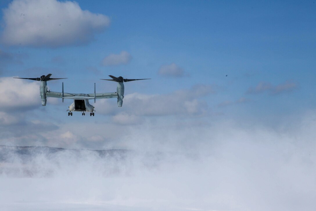 A military aircraft flies in a cloudy sky; mist seen below.