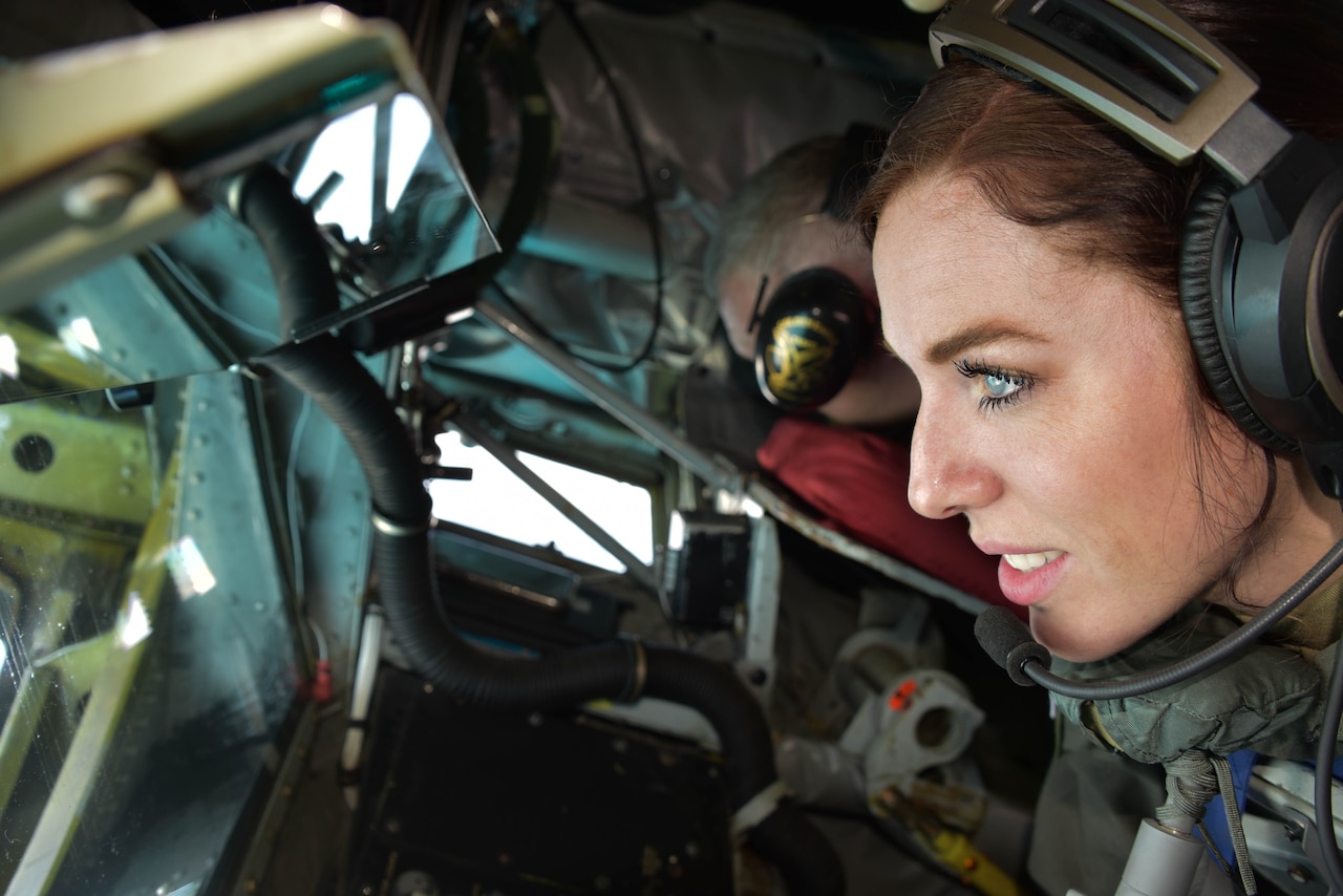 An airman  wearing a headset looks out the window of an aircraft.