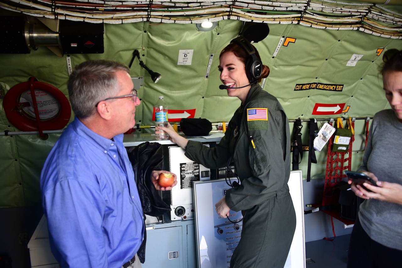 A man speaks to an airman wearing a headset as they stand surrounded by flight equipment.