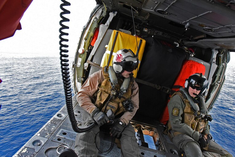 Two sailors sit in a helicopter while traveling over the sea.