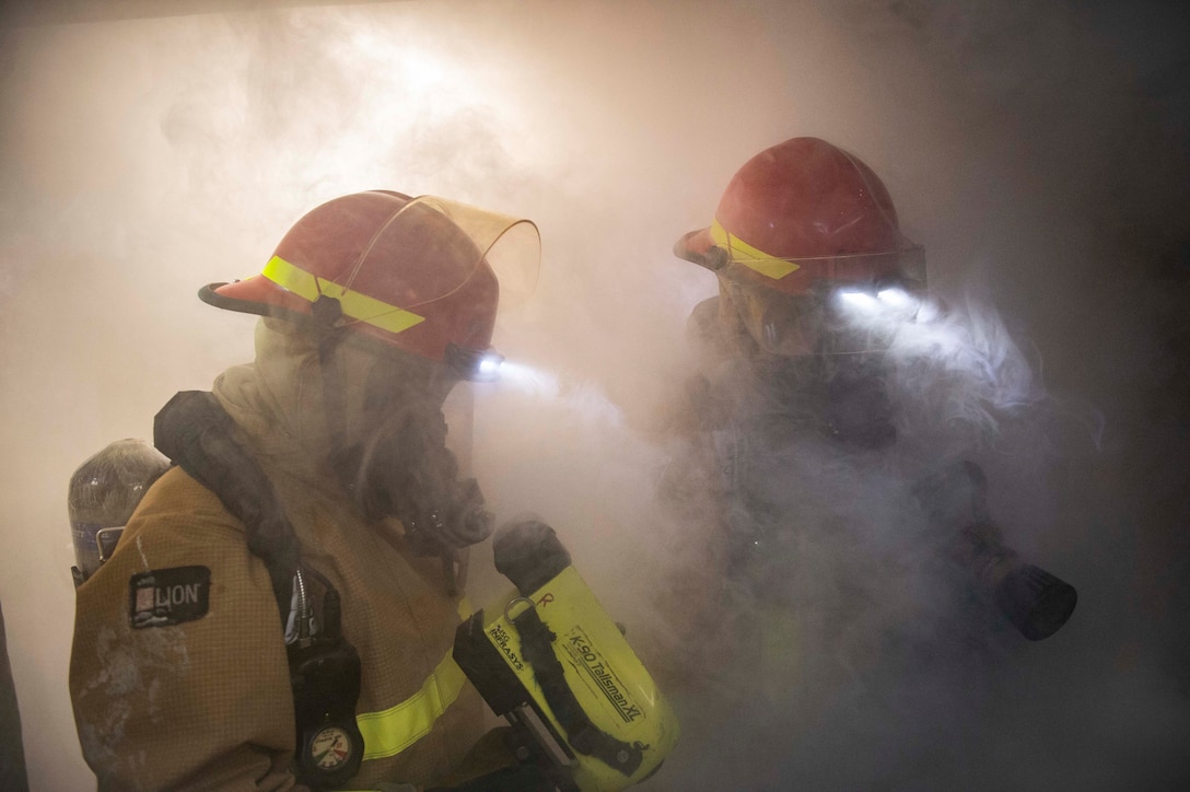Two sailors in fire gear stand in a room filled with smoke.
