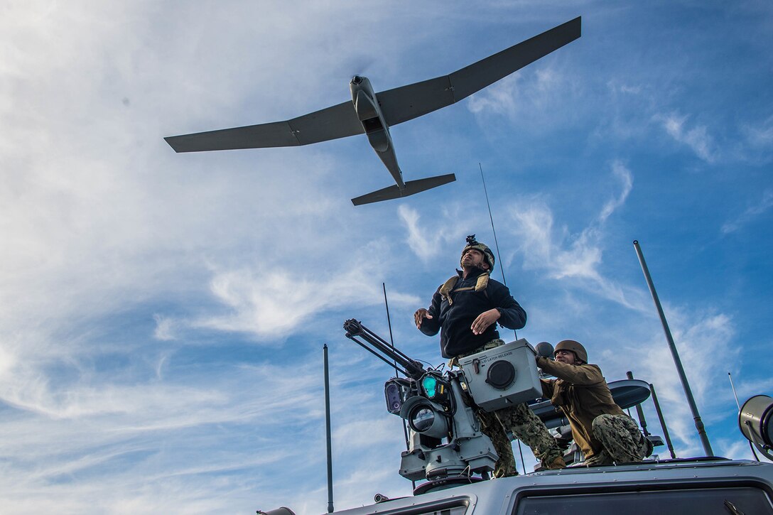 A Navy officer stands on a boat as an unmanned aerial vehicle takes off.