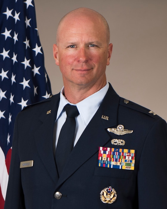 A Caucasian bald male posing for a portrait in front of an American flag, wearing a US Air Force service dress uniform