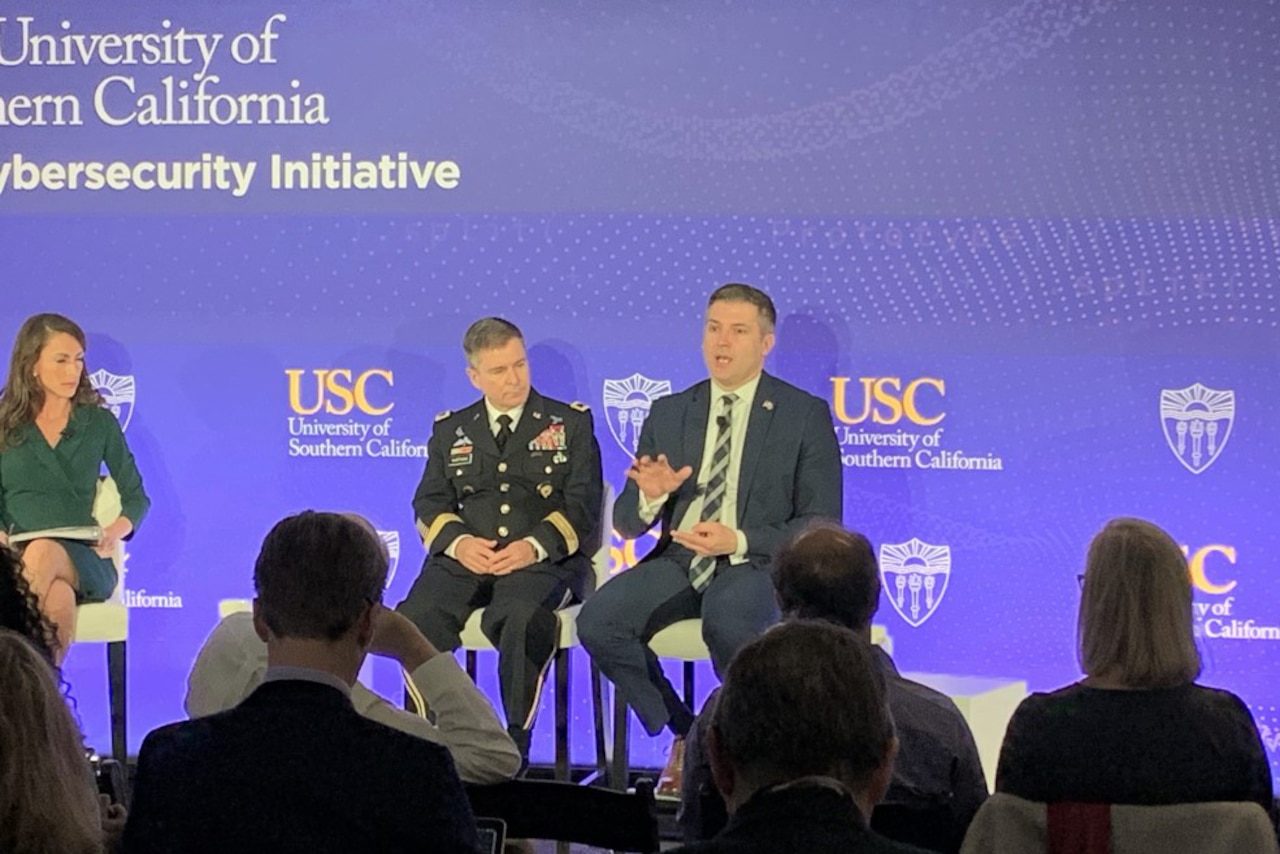 An Army officer and a civilian are seated on a stage facing an audience.