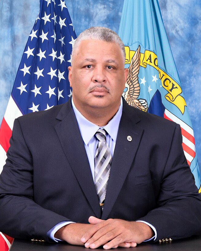 official photo with blue background and American and DLA flags behind man in suit
