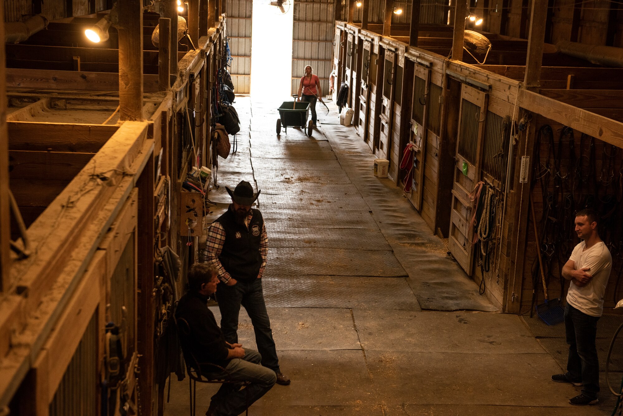 Image of the inside of a horse barn with horse owners talking and doing work.