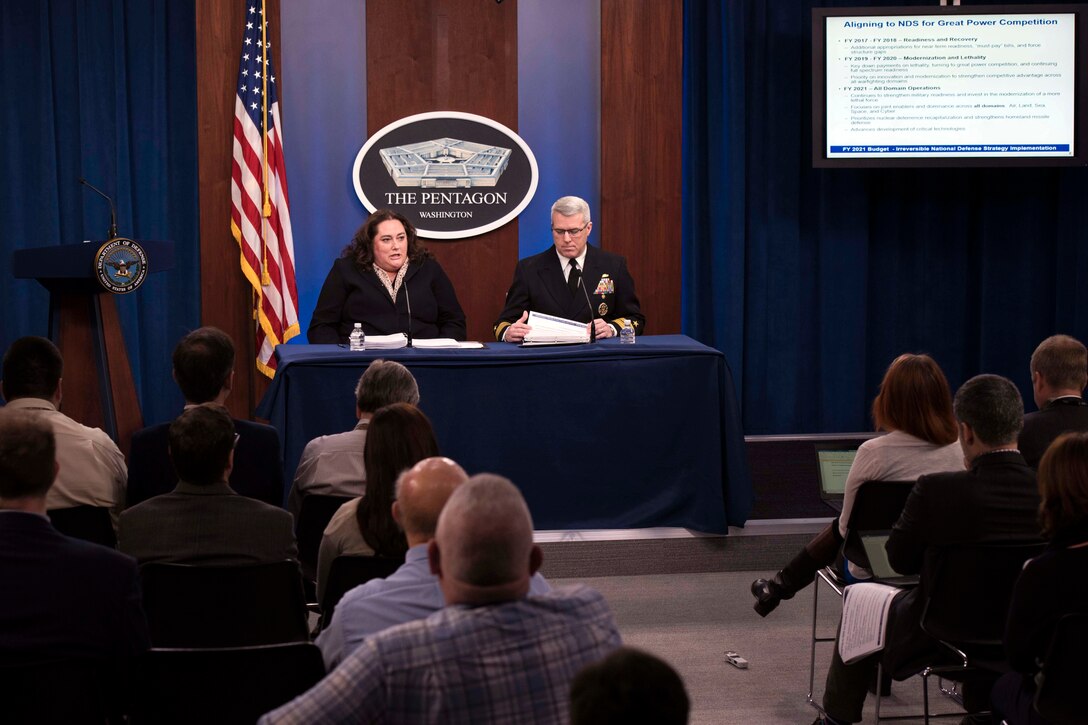 Two people, one civilian and one military, sit behind a desk and talk to reporters.