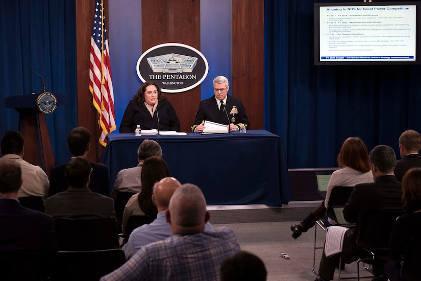 Two people, one civilian and one military, sit behind a desk and talk to reporters.