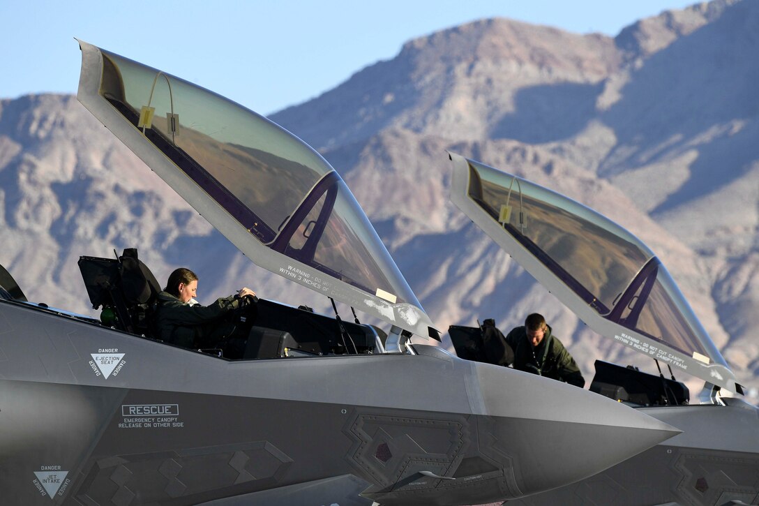 Two airmen sit inside of two parked military jets.