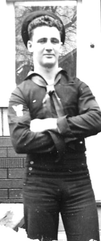 A sailor in his blue working uniform stands with his hands crossed over his chest.