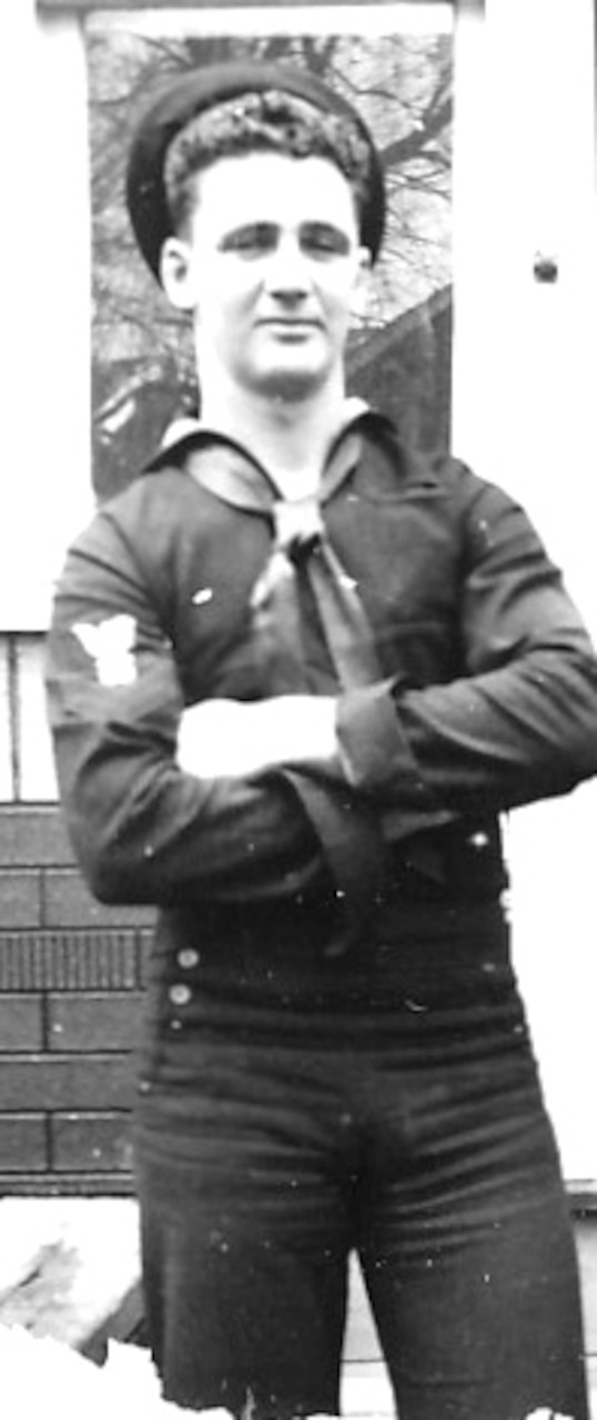 A sailor in his blue working uniform stands with his hands crossed over his chest.