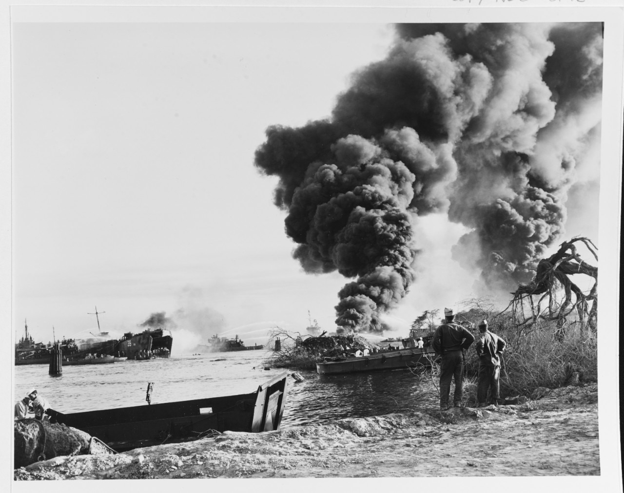 Smoke billows into the sky from two exploded ships that aren’t visible as two men on the shoreline watch. Other ships seen in the distance  fight the fires.