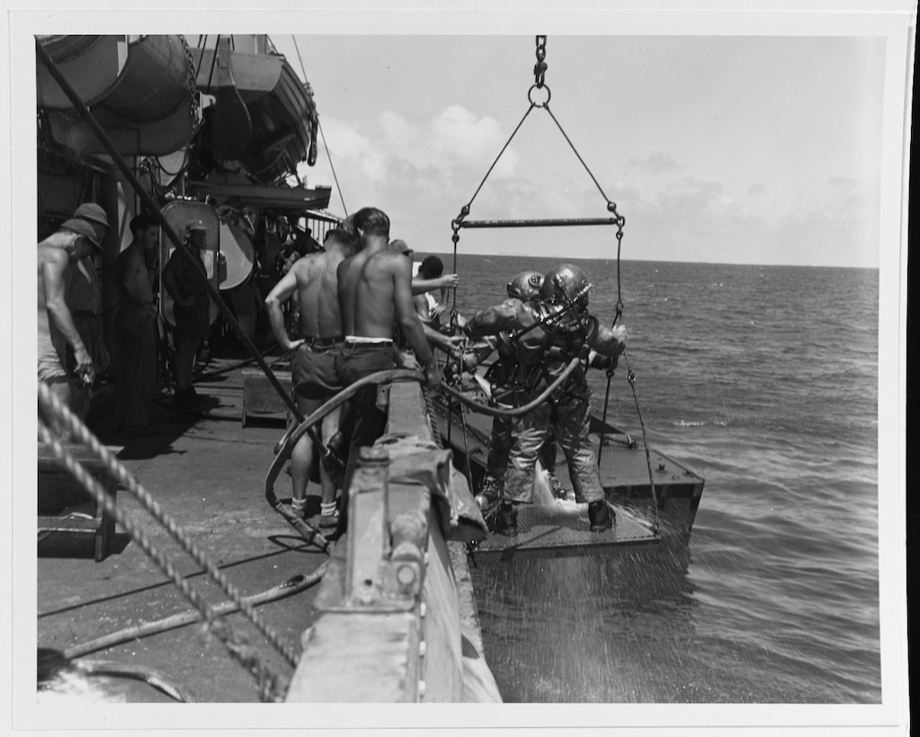 Two men in 1940s-era dive gear stand on a ship’s lift, prepared to be lowered into the ocean.