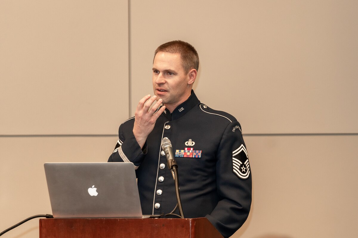 A man who is standing and is dressed in the dark blue Air Force ceremonial uniform is holding his right hand up to his mouth gesturing as he speaks. There is a silver laptop on the brown lectern in front of him.