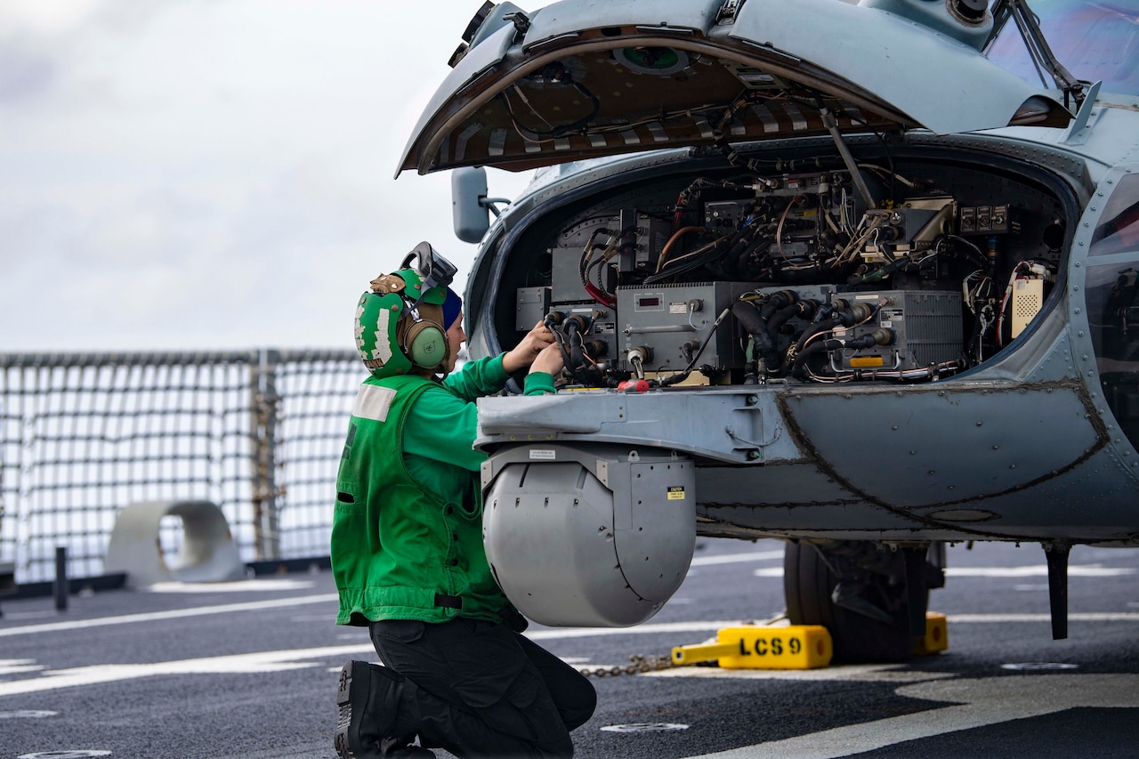 A sailor kneels on a ship's flight deck while working on an aircraft.