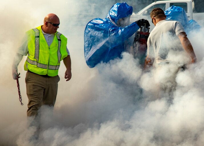 Soldiers stand in a smoky area wearing green and blue protective gear.