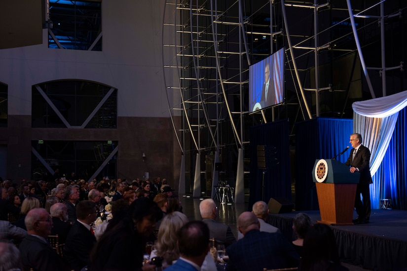 Man speaks at lectern as an audience seated at round tables listens.