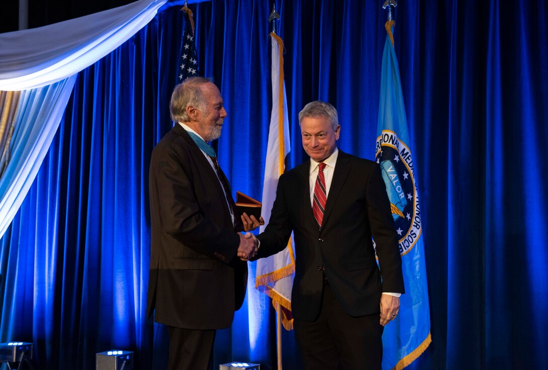 Two men in suits shake hands. One holds an award to present to the other.